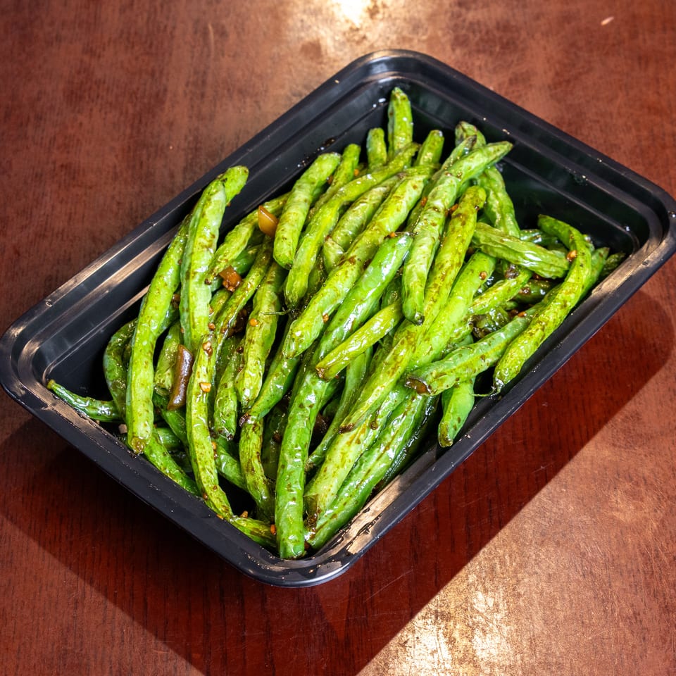 Sautéed green beans with garlic and chili flakes in a black takeout container on a wooden table.