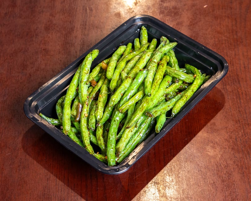 Sautéed green beans with garlic and chili flakes in a black takeout container on a wooden table.