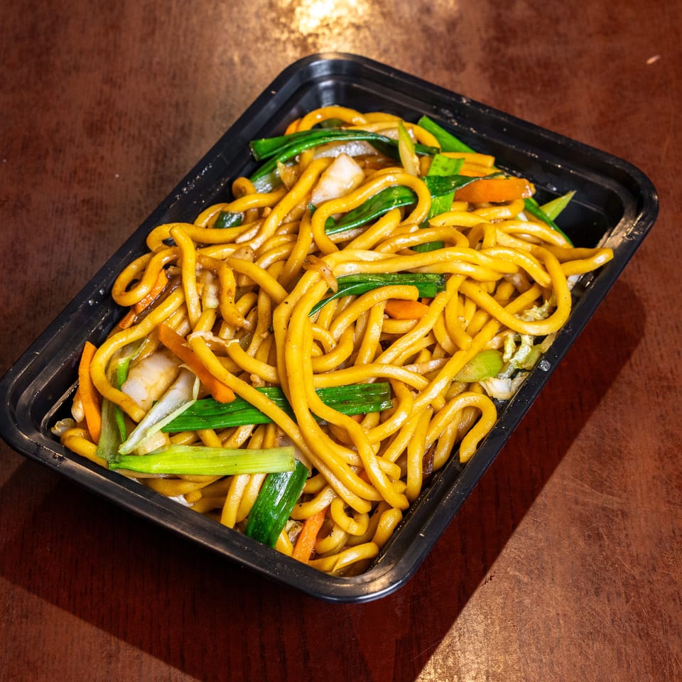 Lo mein noodles with green onions, carrots, and cabbage in a black takeout container.