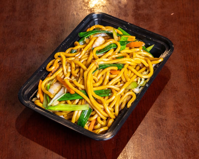 Lo mein noodles with green onions, carrots, and cabbage in a black takeout container.