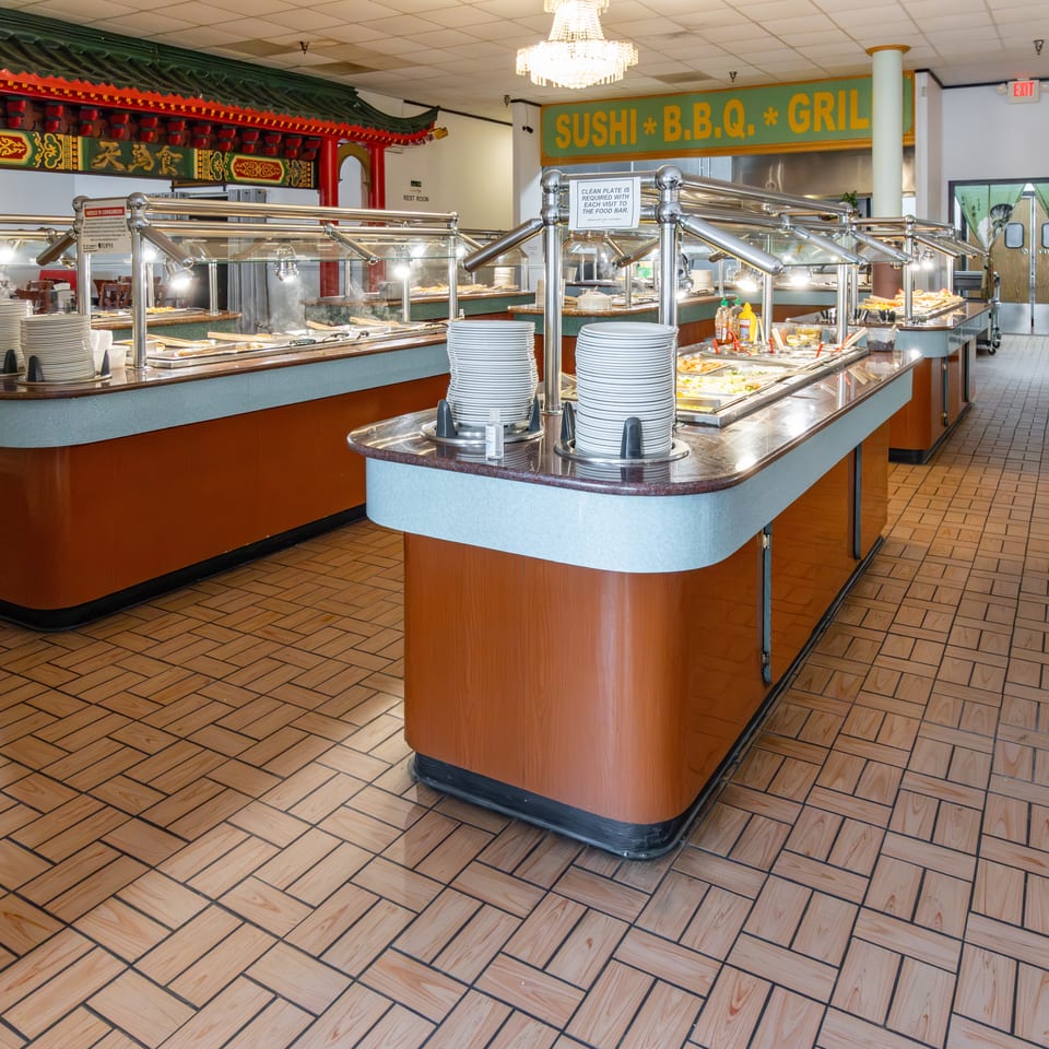 A brightly lit Asian buffet with multiple food stations, stacked plates, and a "Sushi B.B.Q. Grill" sign.