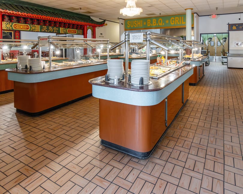 A brightly lit Asian buffet with multiple food stations, stacked plates, and a "Sushi B.B.Q. Grill" sign.