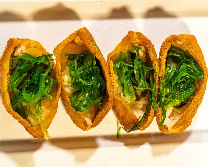 Four golden-brown inari sushi pockets filled with white rice and vibrant green seaweed salad.