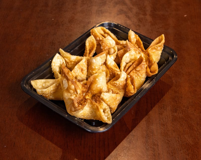 A black rectangular container filled with golden-brown fried wontons, resting on a dark wooden table.