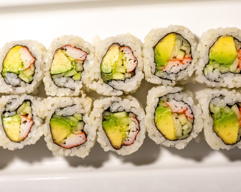 Two rows of California sushi rolls with avocado, cucumber, and crab meat on a white plate.