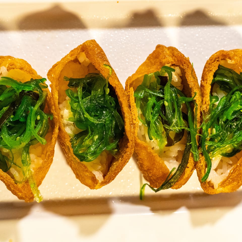 Four inari sushi pieces with rice and bright green seaweed salad on a white plate.
