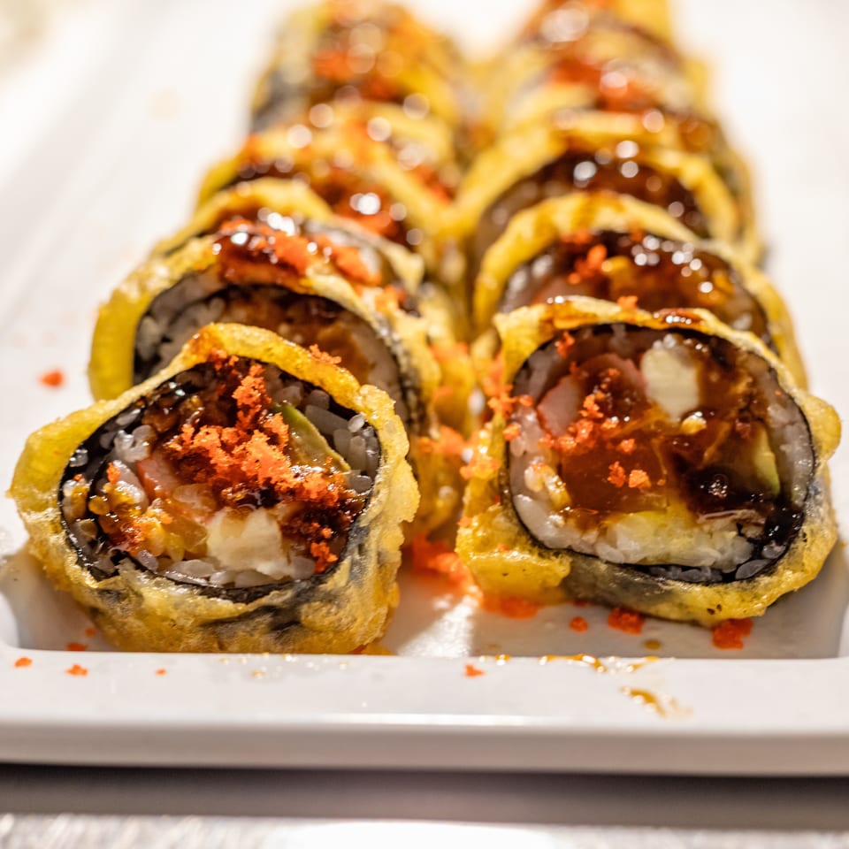 Close-up of a row of deep-fried sushi rolls topped with orange roe and dark sauce.