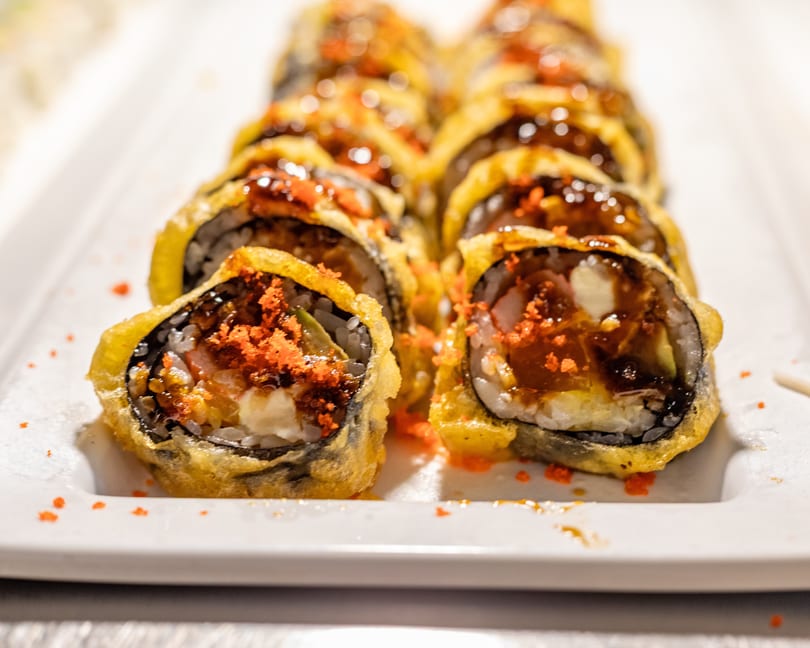 Close-up of a row of deep-fried sushi rolls topped with orange roe and dark sauce.