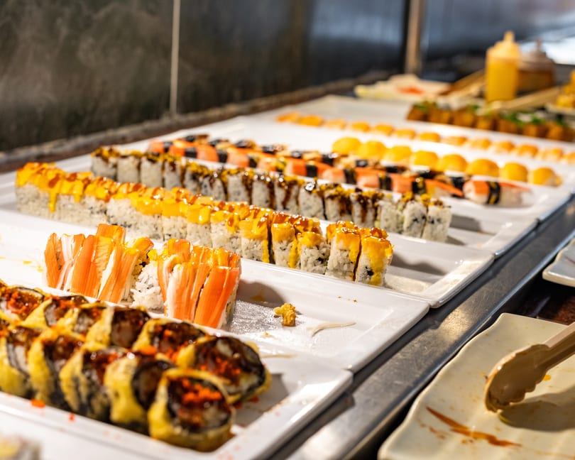 A sushi buffet line with various rolls, including crab, spicy tuna, and fried options.