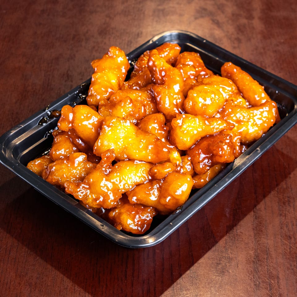 Orange chicken in a black plastic takeout container on a dark wooden table.