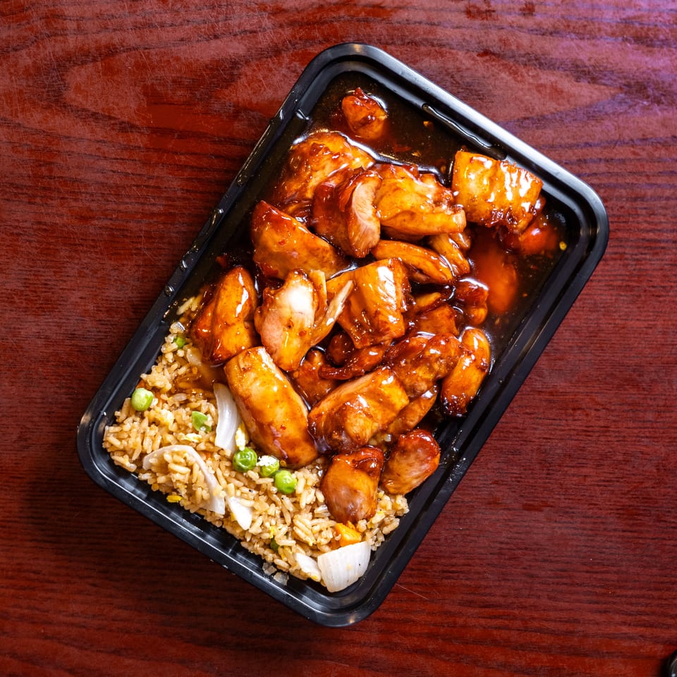 Teriyaki chicken and fried rice with peas and onions in a black takeout container on a wooden table.