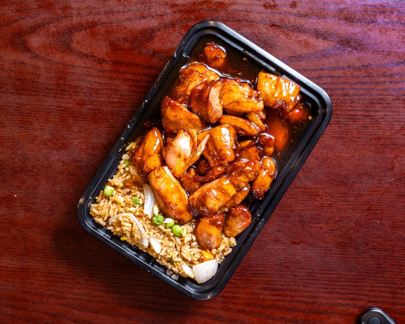 Teriyaki chicken and fried rice with peas and onions in a black takeout container on a wooden table.