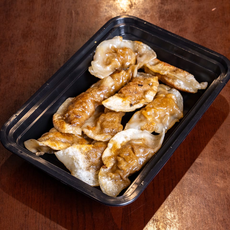 Pan-fried dumplings with a golden-brown crisp, served in a black takeout container on a wooden table.