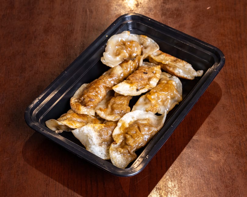 Pan-fried dumplings with a golden-brown crisp, served in a black takeout container on a wooden table.