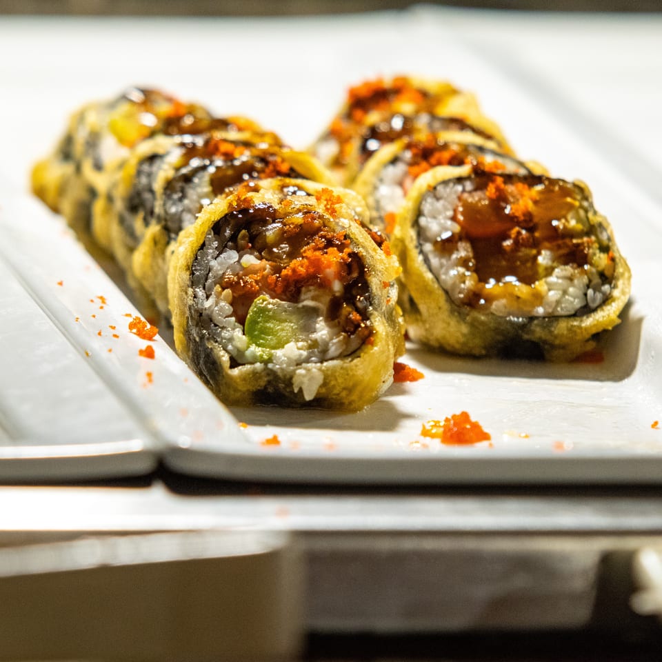 Close-up of fried sushi rolls with avocado, topped with sauce and orange roe on a white plate.