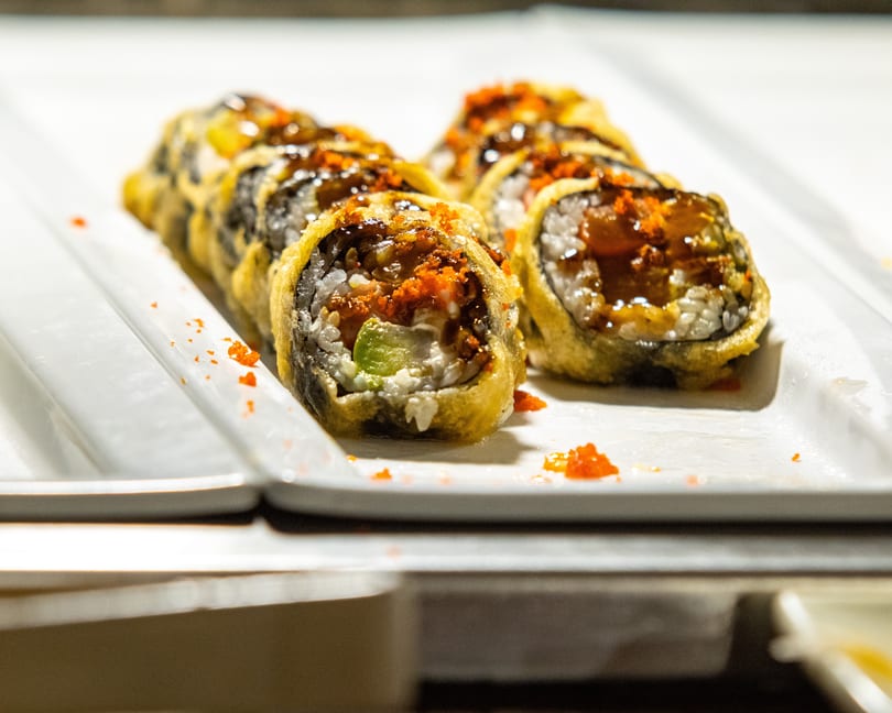 Close-up of fried sushi rolls with avocado, topped with sauce and orange roe on a white plate.