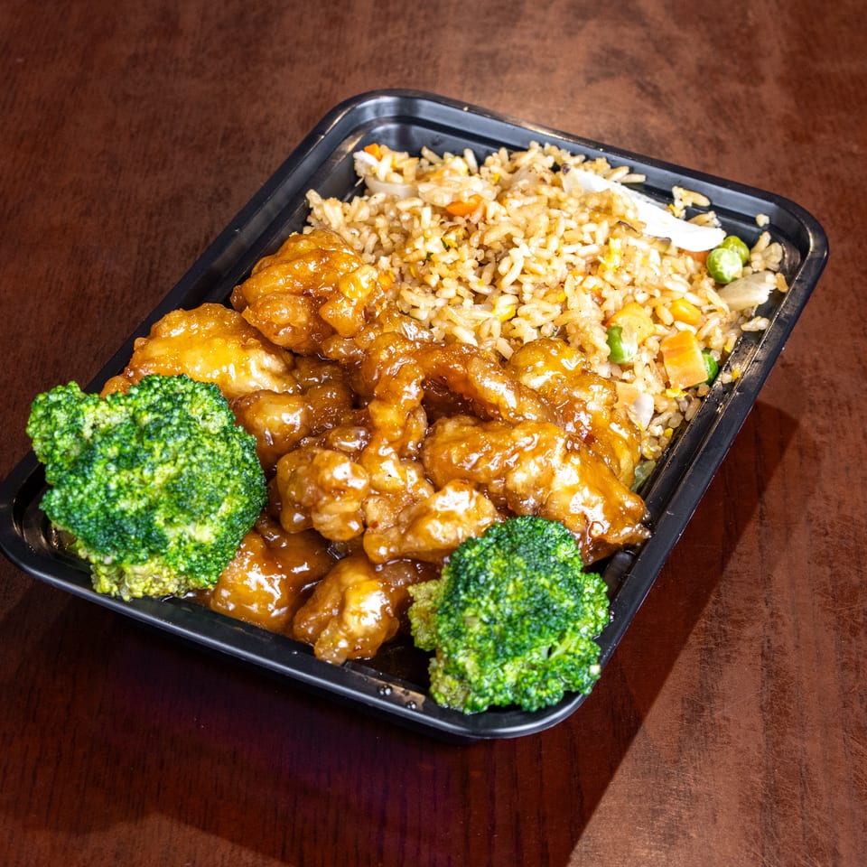 Orange chicken with fried rice and broccoli in a black takeout container on a wooden table.