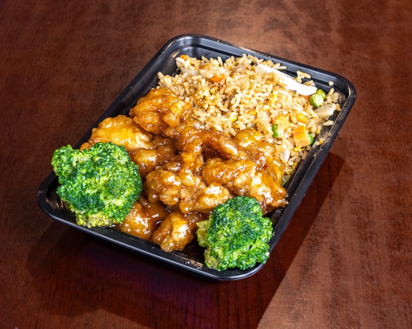 Orange chicken with fried rice and broccoli in a black takeout container on a wooden table.
